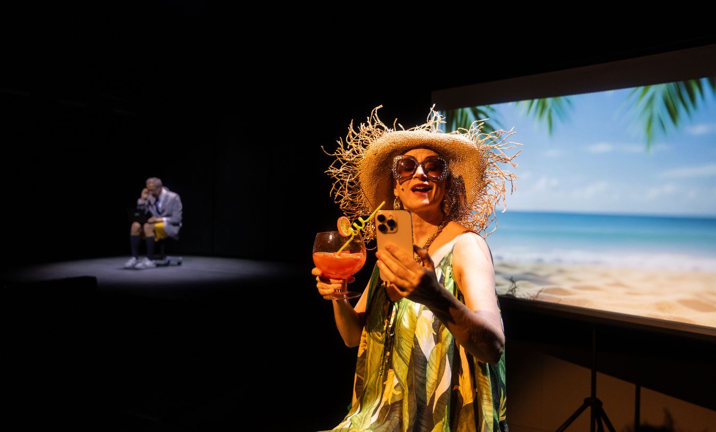 Skodespelar Christine Hope i strandkjole med solbriller og drink snakkar med lærar Thomas Bye som sit på pult i bakgrunnen.