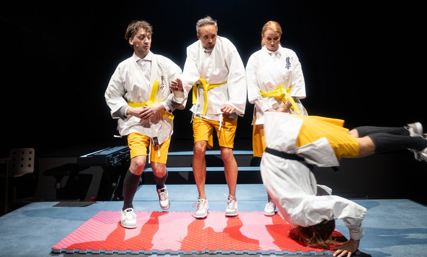 Fire skodespelarar i gule shorts og kvite judodrakter står på ei matte. Ein av dei er i lufta med hovudet ned i salto.