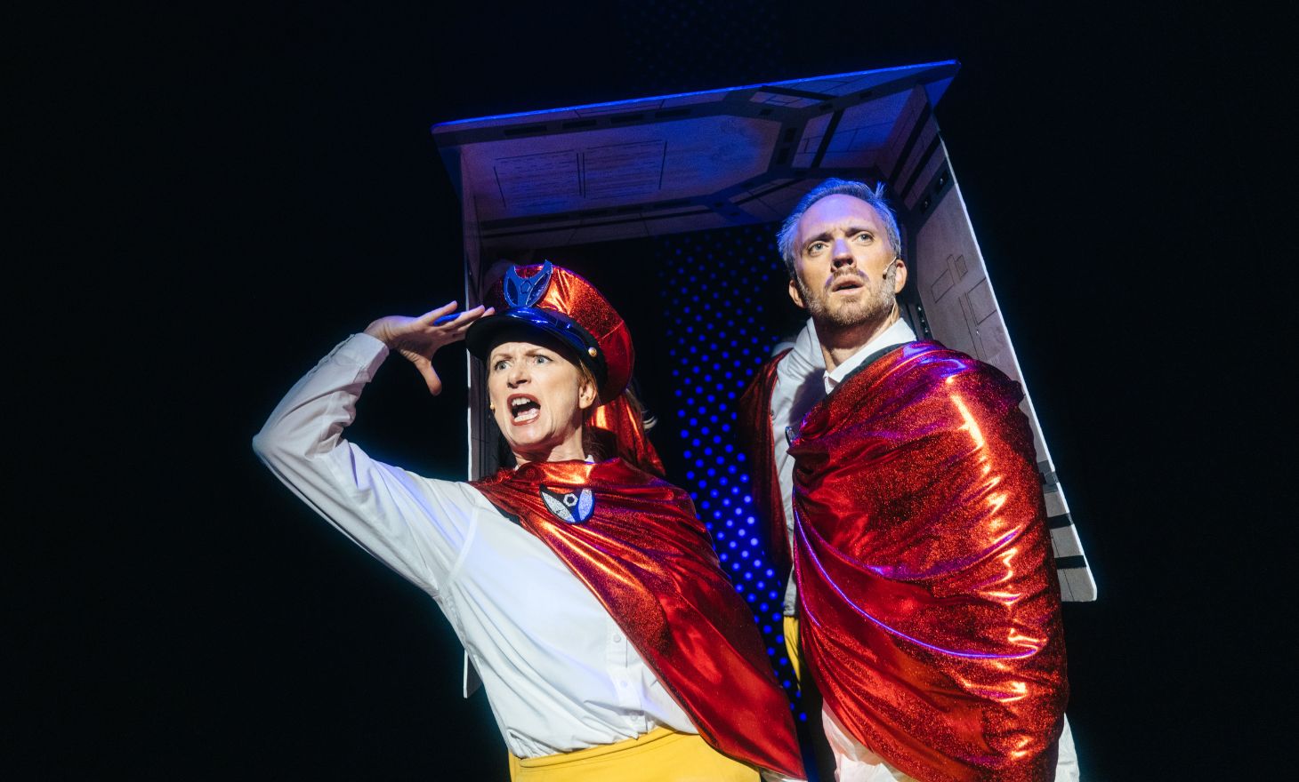 Skodespelarar i raude kapper og fantasifulle kostyme ser dramatisk ut i spotlight på scena.