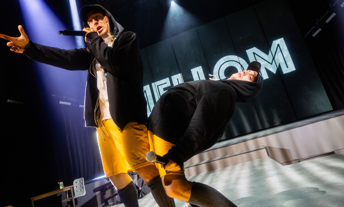 To skodespelarar i hettegenser og caps står på scena med mikrofon og rappande uttrykk i sterkt scenelys.